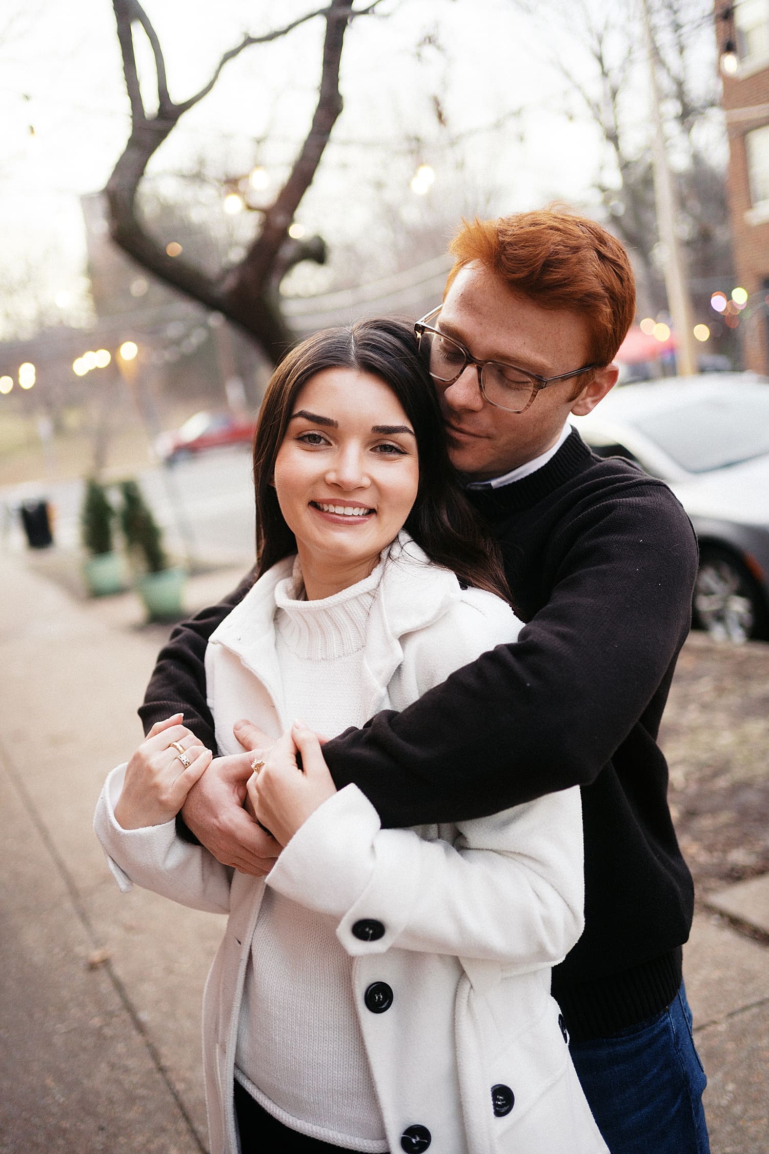 brunette in white coat is hugged by redhead on sidewalk showing your engagement session should happen where you live