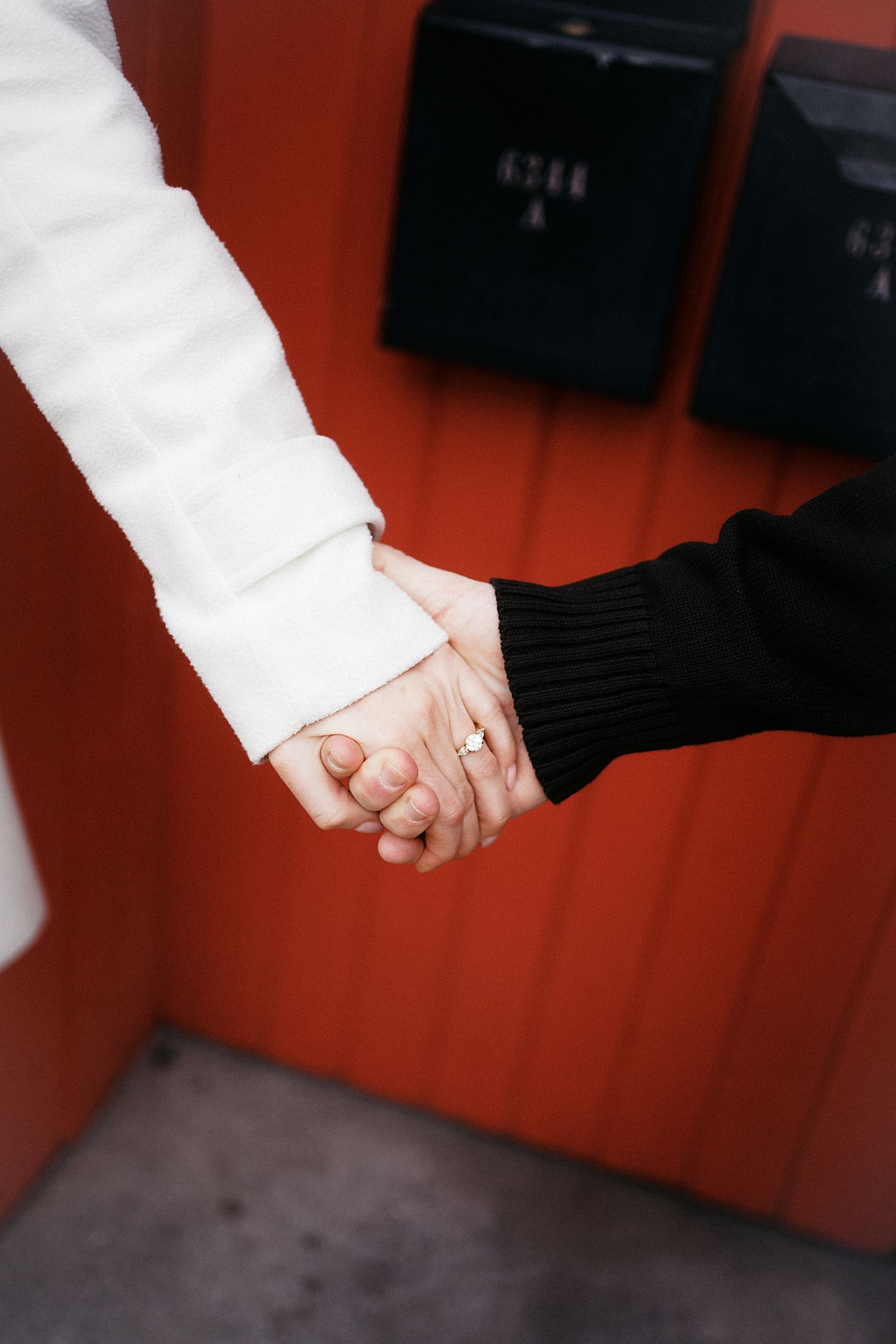 two people hold hands in front of red wall with diamond ring on finger by Camille Meers Photography