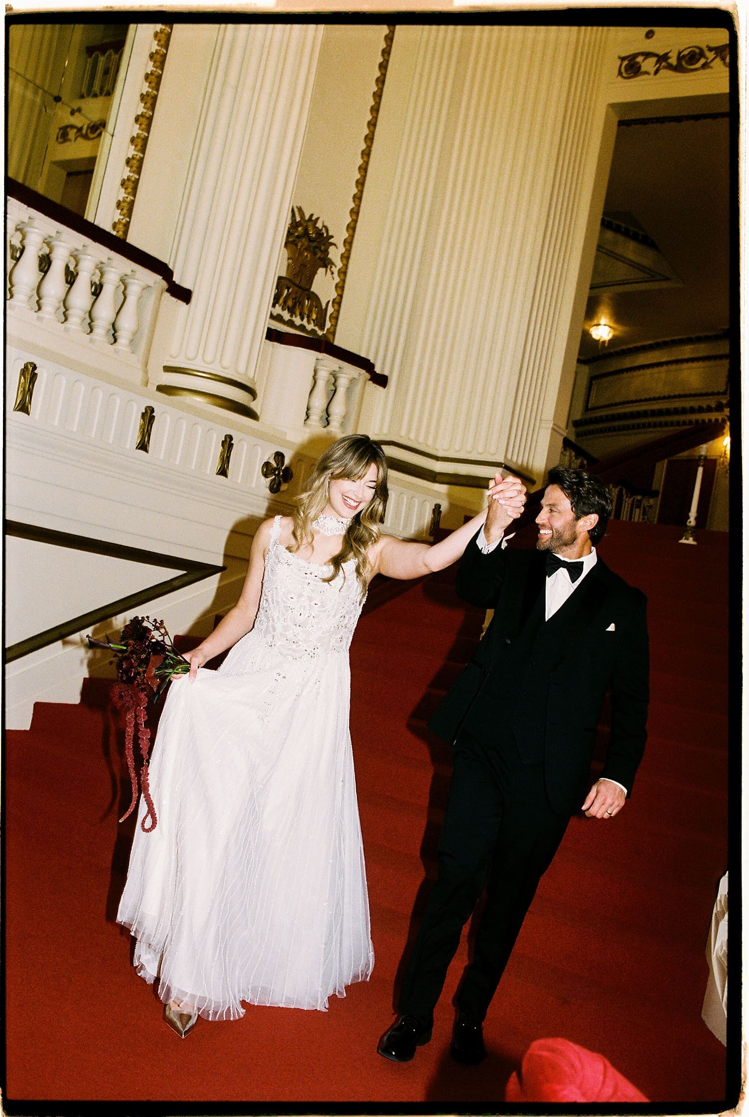 newlyweds walk down grand red staircase by St. Louis wedding photographer