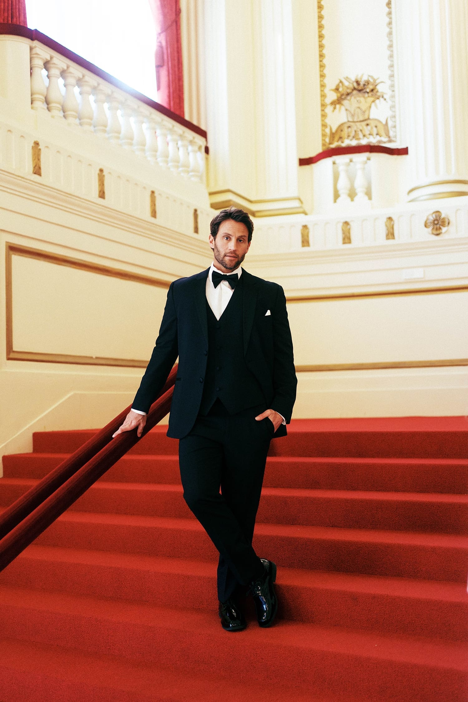 groom in tux leans on railing of red stairs by St. Louis wedding photographer