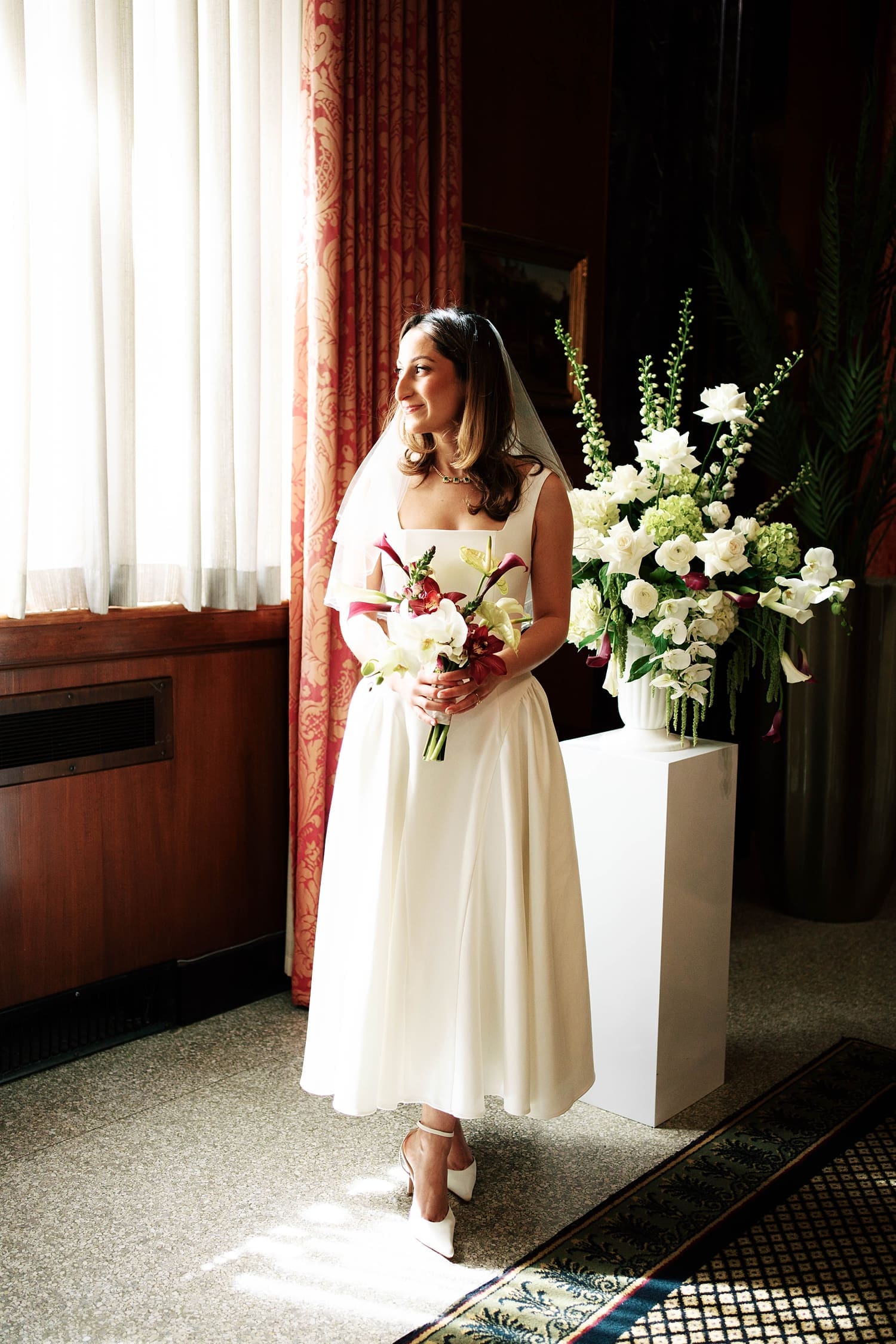 bride in midi dress stands in front of window at the Missouri Athletic Club