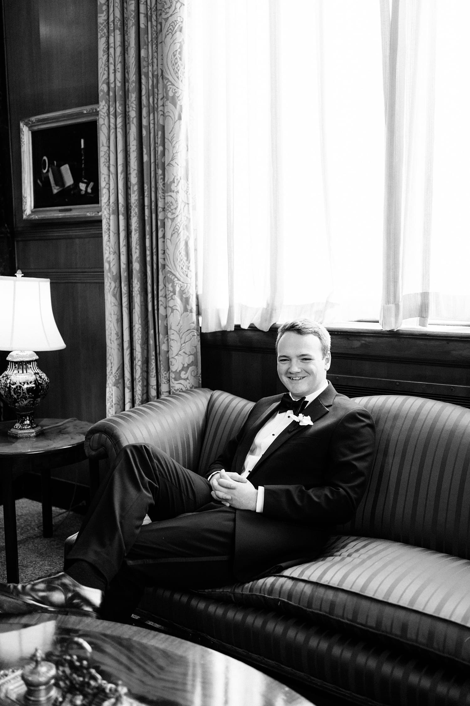 groom sits on vintage coutch in front of large window by Camille Meers Photography