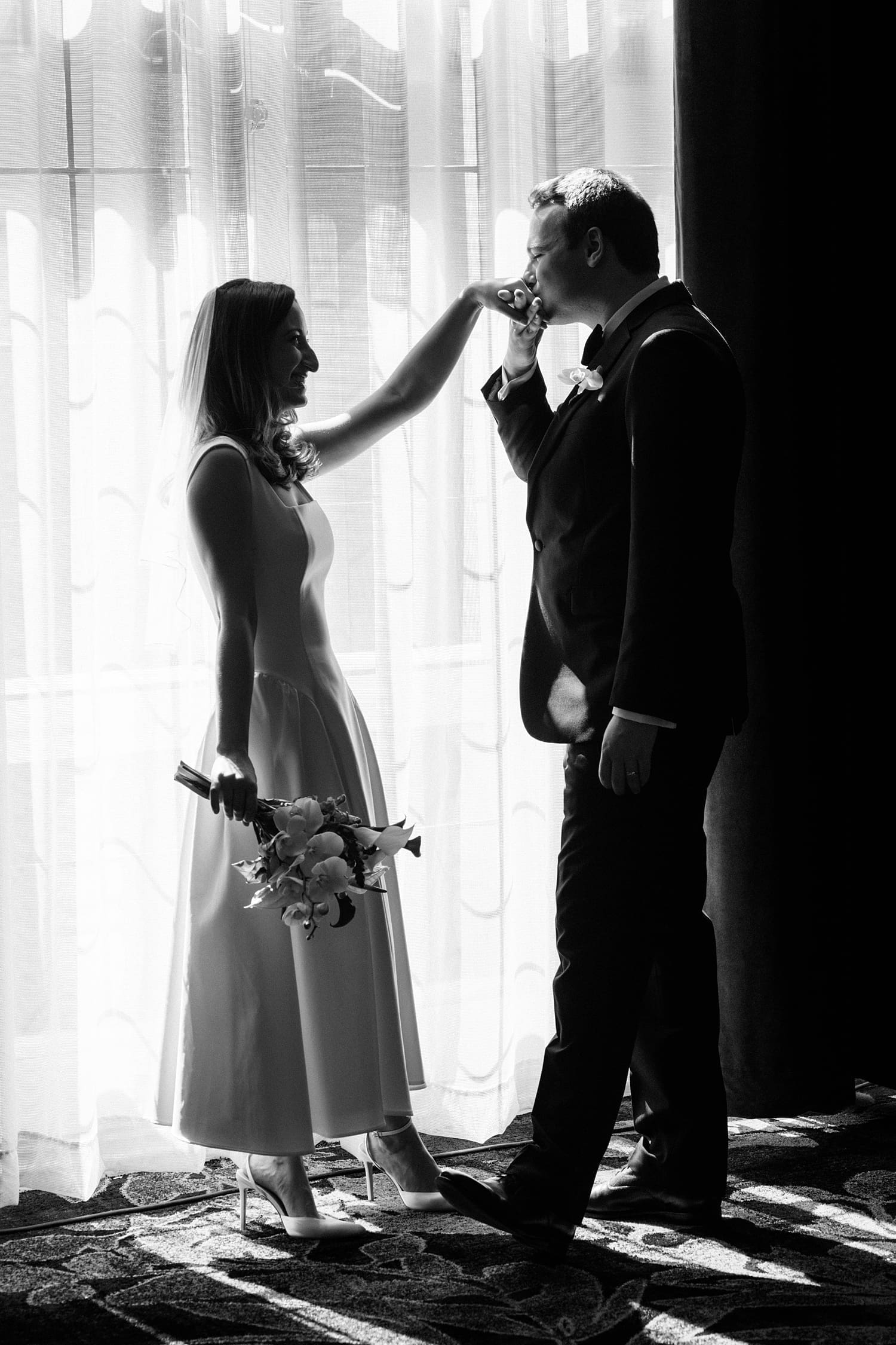 groom kisses his bride's hand in front of a window at the Missouri Athletic Club