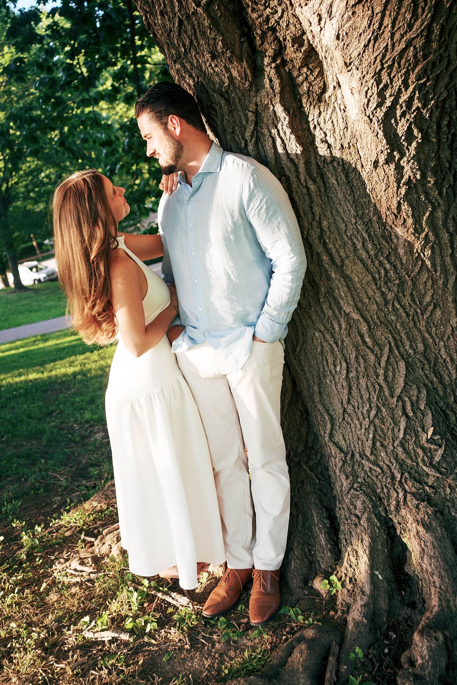 brunette woman in white dress leans up against her man by tree by documentary wedding photographer