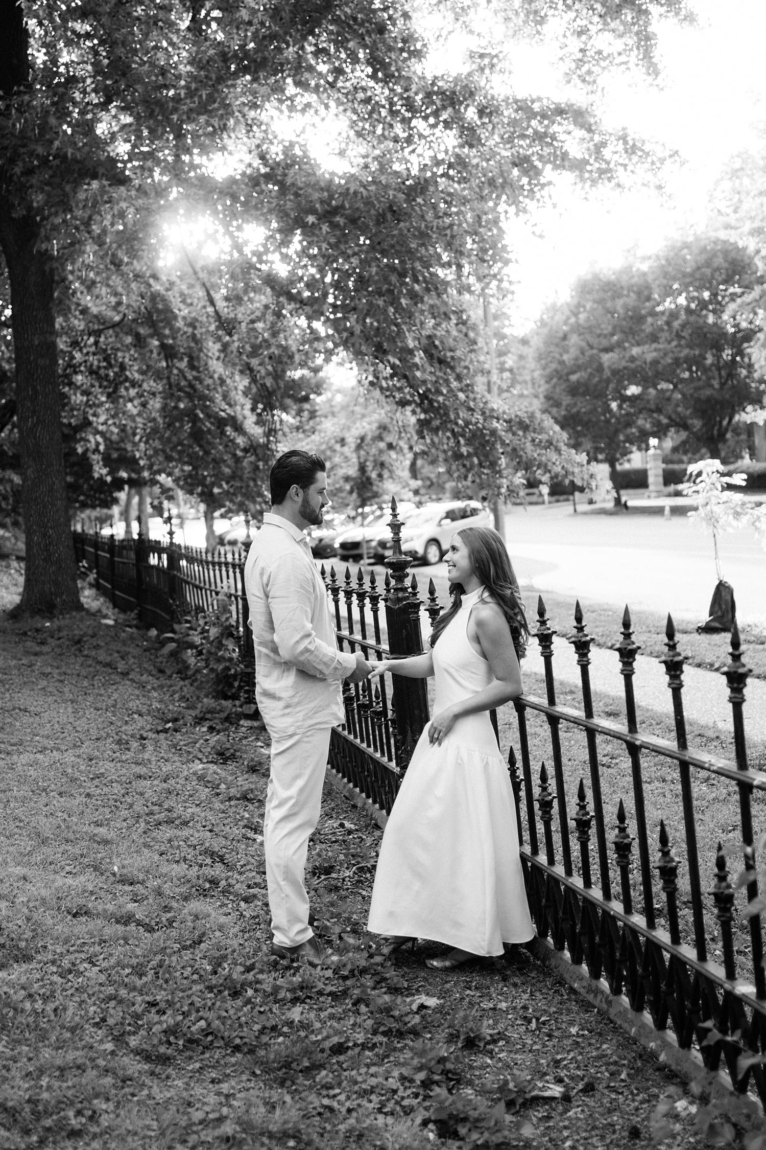 woman leans against fence in park while her fiancé holds her hand by Camille Meers Photography