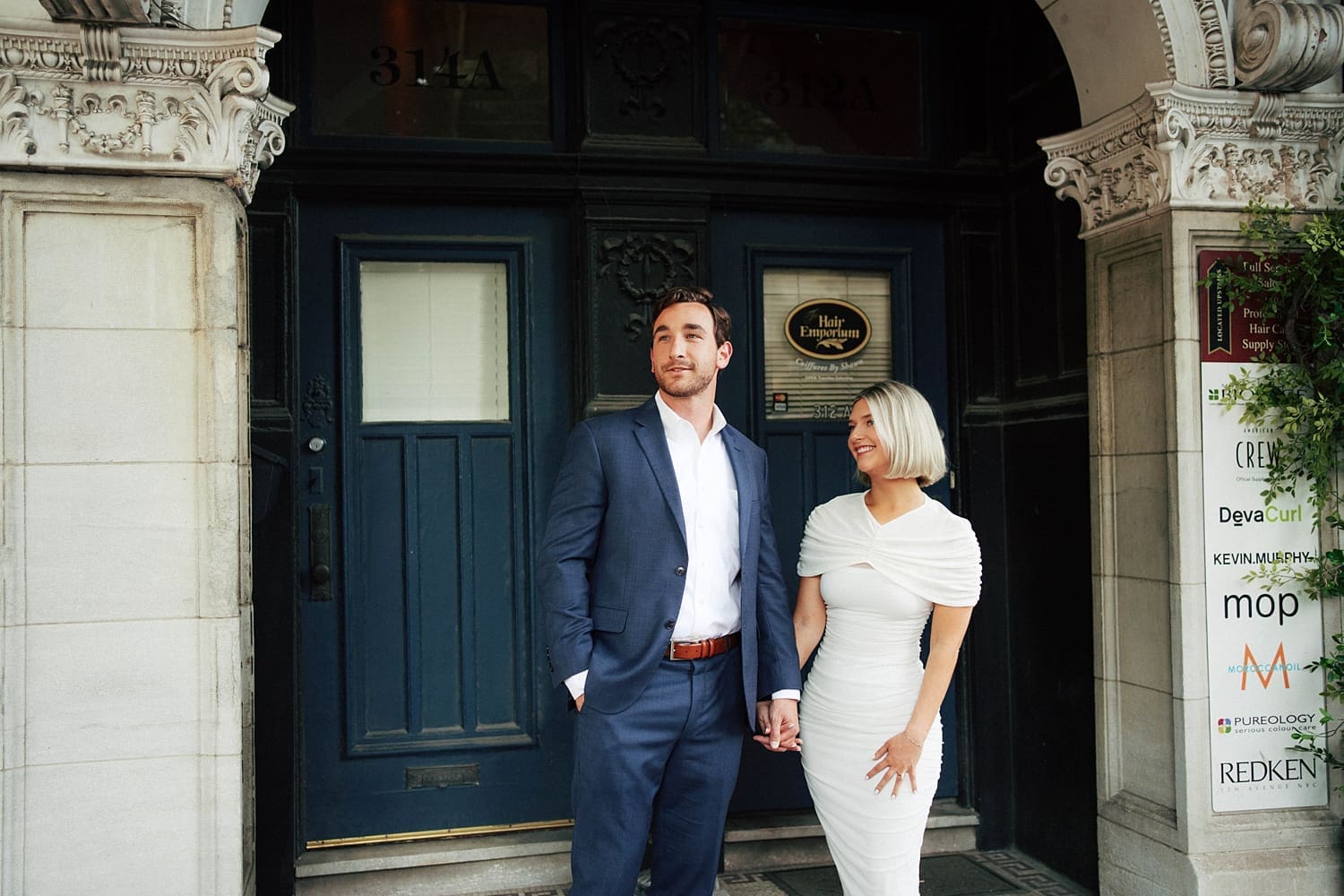 man in blue suit and woman in white dress stand outside restaurant by Camille Meers Photography